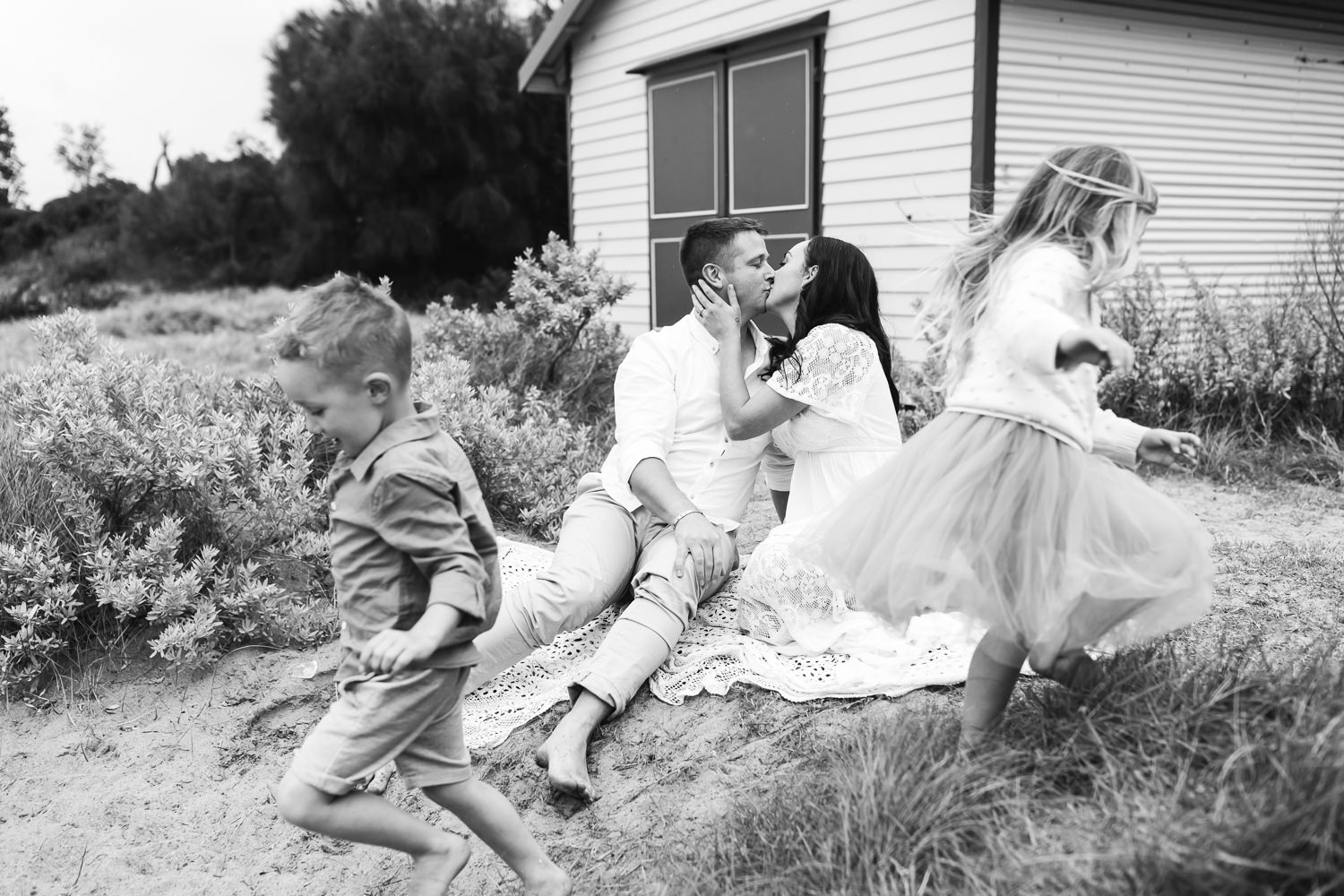 A couple sits on the dunes kissing while their two children run around them playfully.