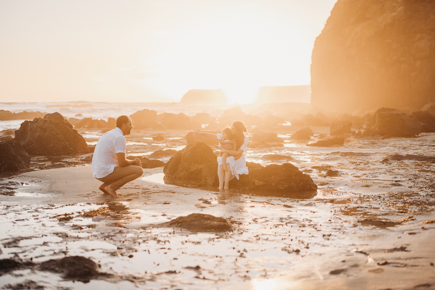 A mother holds her toddler while sitting on a rock at the beach. A man crouches next to them as the sunsets over a cliff in the background.