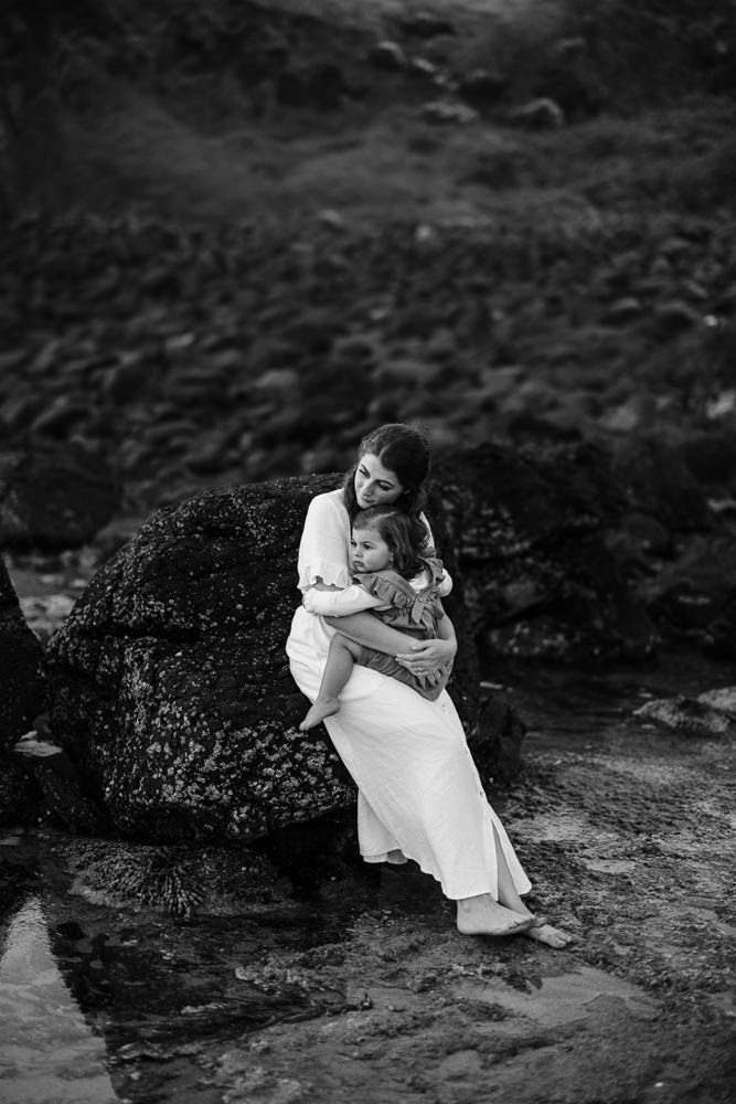 A mother sits on a rock cradling her daughter. Her eyes are closed. Black and white photograph