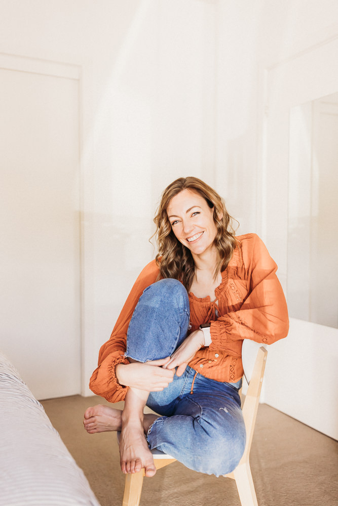 Merissa sits on a chair in her bedroom. Her hands wrap around her knee and she smiles at the camera