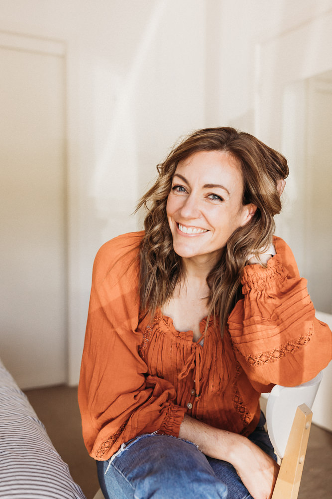Merissa sits smiling at the camera. Her left hand scrunches her hair and she's wearing an orange boho style top.