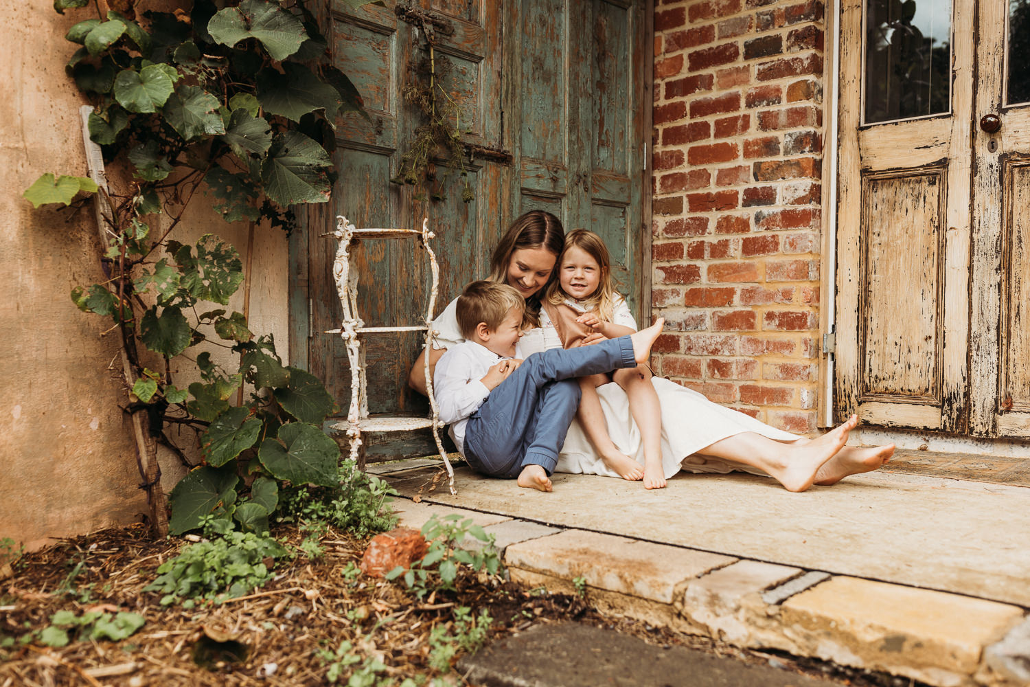 A mother and her two children sit in a corner of the garden at the potterage in Menzies Creek in the Dandenong Ranges. They lean on a vintage green wooden door.
