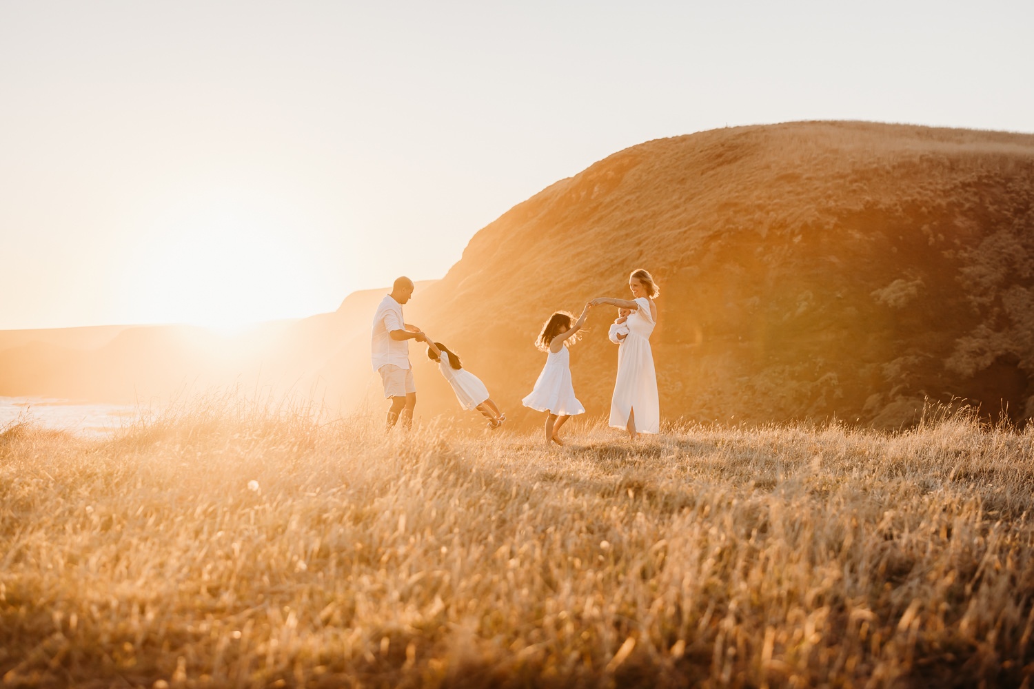 A family of five stand on a golden cliff at sunset. The father swings on of his daughters around as another daughter twirls in the golden light. The mother holds a baby girl and looks at her family.
